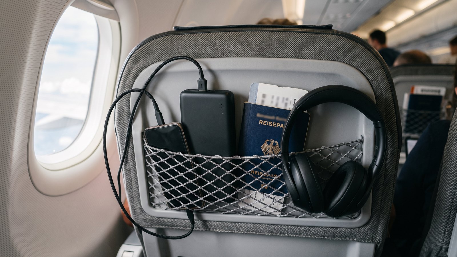 Powerbank mit angestecktem Kabel in der Netztasche an der Rückenlehne eines Flugzeugsitzes, daneben Reisepass und Kopfhörer, Tageslicht vom Kabinenfenster