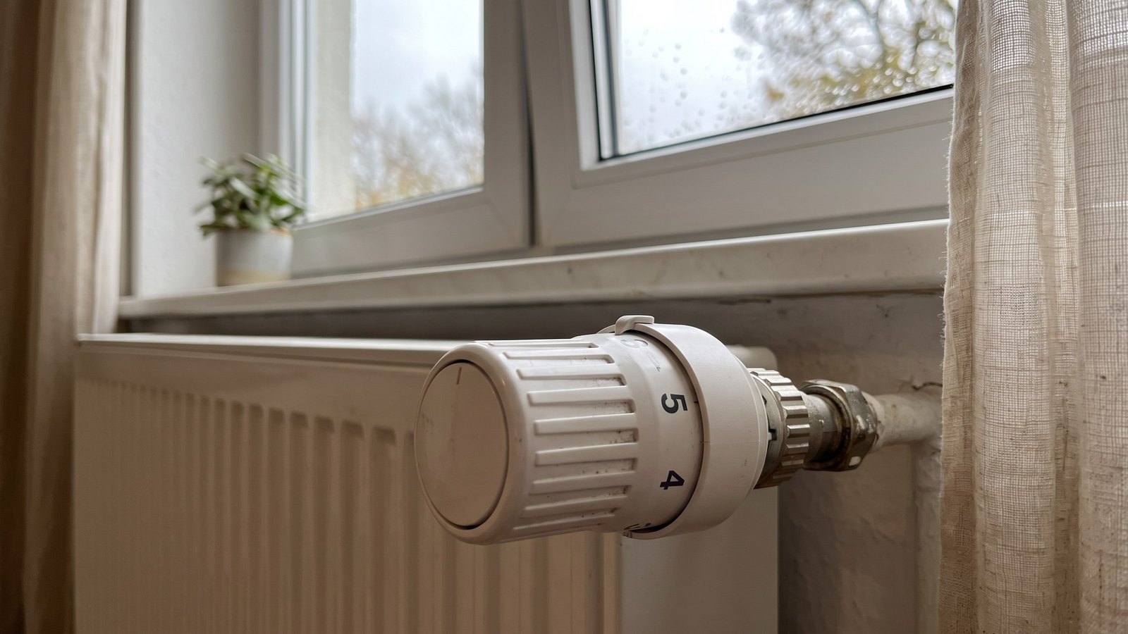 Weißer Heizkörperthermostat mit Drehknopf an einem Heizkörper unter einem Fenster, die Skala zeigt die Ziffern 4 und 5